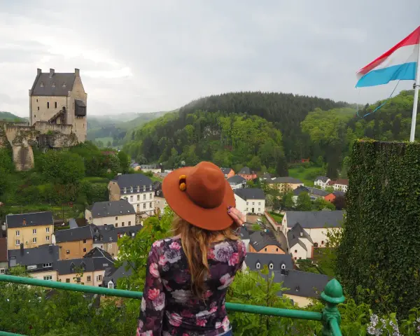 Mujer con sombrero marrón admira un castillo y un pueblo, con la bandera de Luxemburgo ondeando. ¡Un paisaje digno de un buen queso Muenster!