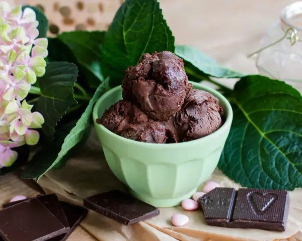 Ein grüner Napf mit zartbitter schokoladeneis, umgeben von Schokoladentafeln, rosa Bonbons und einer Hortensienblüte.