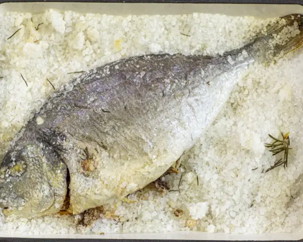 Orata al sale, intera e dorata, adagiata su un letto di sale grosso e rosmarino in una teglia rossa.
