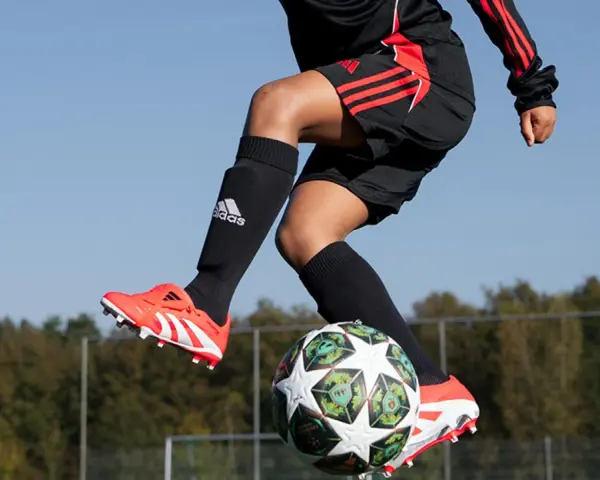 Barn i luften med fotboll. Svarta strumpor med Adidas-logga och benskydd fotboll barn storlek. Orange fotbollsskor med vita detaljer.