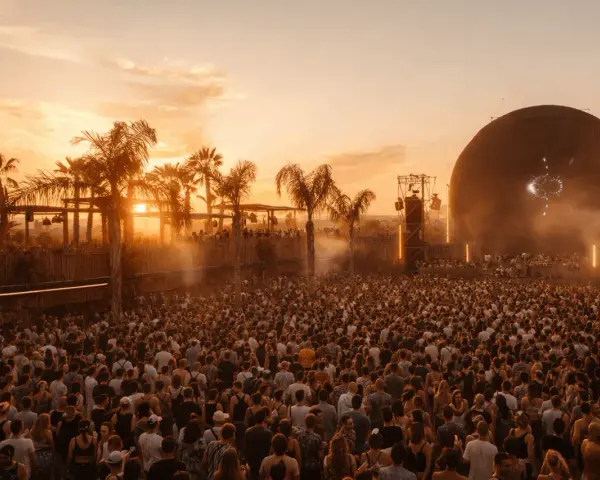 Multitud en el festival Sophie, disfrutando del atardecer con palmeras y una gran esfera como escenario.
