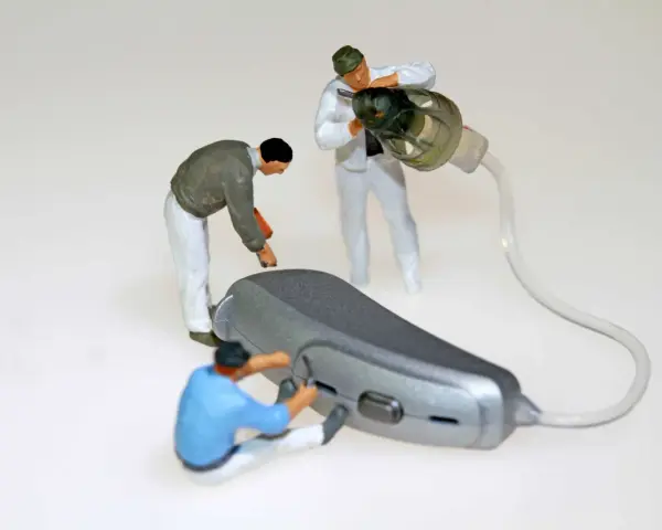 Miniaturowi pracownicy zajmują się naprawą aparatów słuchowych. Jeden z nich przykręca element, drugi czyści słuchawkę, a trzeci pracuje nad aparatem.