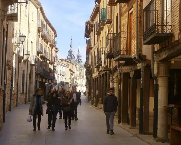 Gente pasea por una calle porticada, un lugar ideal para descubrir qué ver en Burgo de Osma. Edificios históricos y torres se alzan al fondo.
