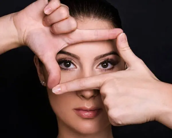 Mani incorniciano il volto di una donna, creando un effetto di profondità. Si può vedere un alone nell'occhio, quasi un riflesso di luce.