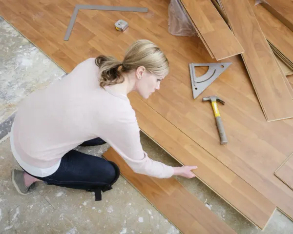 Frau kniet und verlegt Laminat. Werkzeuge wie Hammer und Winkel liegen bereit.