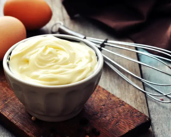 Un bol de mayonnaise onctueuse, des œufs et un fouet, prêts à faire une mayonnaise maison.
