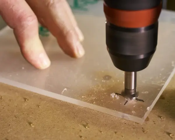 A drill bit is used to drill through acrylic. A hand guides the acrylic sheet as the drill creates a hole, showing it's possible to drill through acrylic.