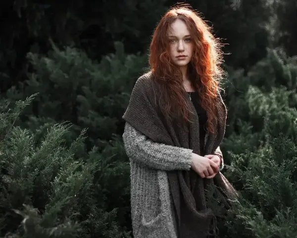 Rudowłosa dziewczyna w grubym swetrze i szalu, otoczona ciemną zielenią. Jej spojrzenie jest spokojne, a cała scena ma **low-key** klimat.
