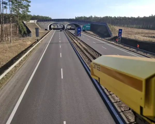 Żółta kamera monitoruje odcinek pomiarowy prędkości na S8. Droga prowadzi przez las, pod wiaduktem.