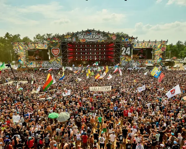 Tłum ludzi na festiwalu Poland Rock, gdzie jest scena i ekrany. Wszędzie flagi i słońce.