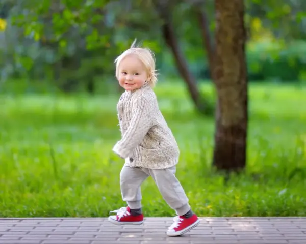 Ein fröhliches Kind, ca. 2,5 Jahre alt, mit blonden Haaren und einem Strickcardigan, läuft auf einem Weg im Park.