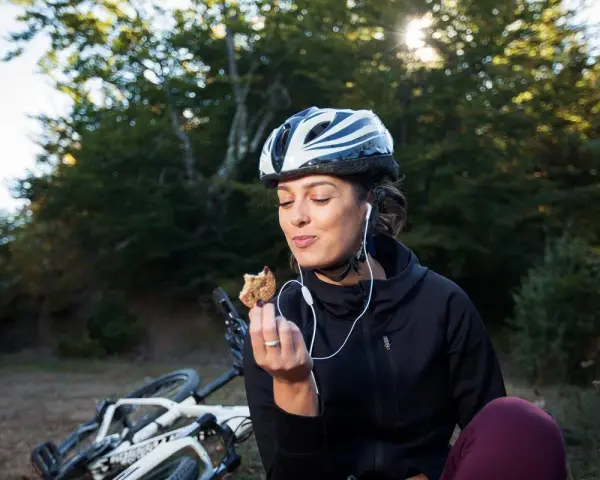 Kobieta w kasku i słuchawkach je ciastko, obok leży rower. To przykład zdrowego odżywiania i aktywnego stylu życia, czyli zbilansowana dieta.