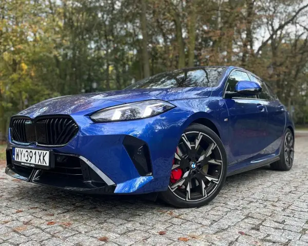 Błękitne, sportowe niemieckie auto BMW z czarnymi felgami i czerwonymi zaciskami hamulcowymi stoi na brukowanej drodze wśród jesiennych drzew.