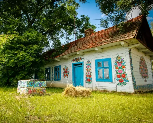 Malowany dom w Zalipiu, z niebieskimi oknami i drzwiami, ozdobiony kwiatami. Obok studnia i snop siana. Cennik wizyty w tym malowniczym miejscu jest dostępny dla zwiedzających.