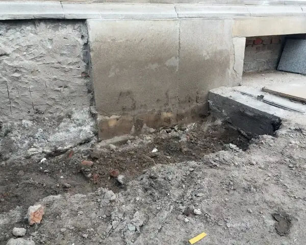 Prace przy wzmocnieniu fundamentów w starym domu. Widoczne wykopy, odsłonięta ściana i nowe elementy betonowe.