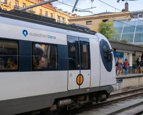 A modern Euskotren train at San Sebastian train station. Passengers wait on the platform under a glass-covered canopy.