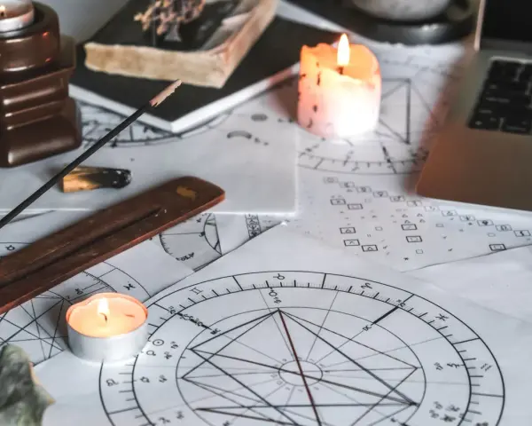 Na stole leżą wykresy astrologiczne, świece, kadzidełko i laptop. Widać symbolikę wskazującą na ascendent w bliźniętach.