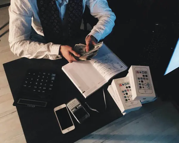 Mężczyzna liczy pieniądze przy biurku z kalkulatorem, notatnikiem, telefonami i pudełkami po iPhone'ach. To może być jego sposób na zarządzanie finansami, być może w kontekście aml.