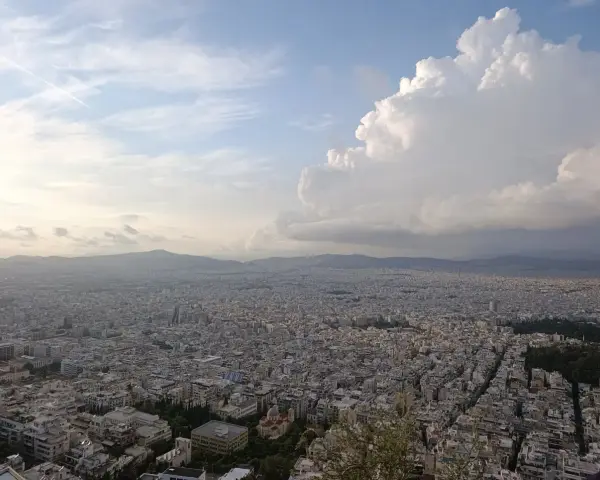 Rozległa panorama Aten z wzgórza Lycabettus. Miasto rozciąga się aż po horyzont, a nad nim piętrzą się chmury.