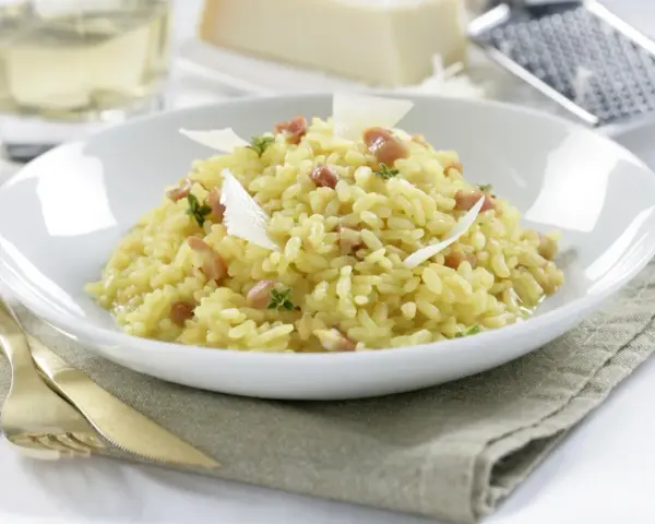 Un cremoso arroz a la milanesa, adornado con láminas de queso parmesano y hierbas frescas, servido en un plato blanco.