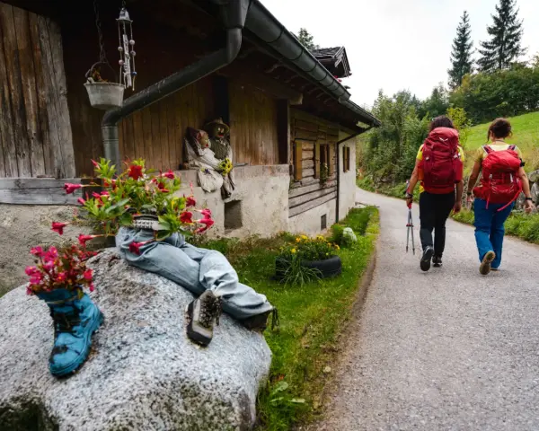 Due escursioniste affrontano il cammino San Vili, con zaini rossi e bastoncini. La strada, tra case rurali e verde, suggerisce una certa difficoltà.