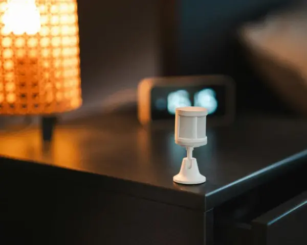 Sensor de movimiento blanco sobre mesita de noche, junto a lámpara y reloj. Ideal para saber como dejar fijo un sensor de movimiento.