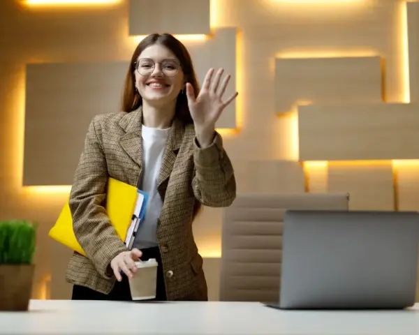 Uśmiechnięta kobieta w okularach macha ręką, trzymając teczkę i kawę. Czy dzień próbny jest płatny?