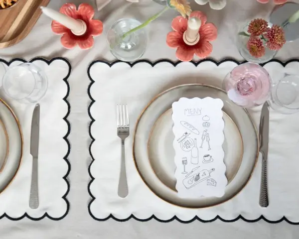 A table setting with a gold-colored tablecloth, white placemats with black scalloped edges, plates, cutlery, glasses, and a menu with illustrations.