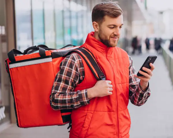 Uśmiechnięty dostawca jedzenia z dużą czerwoną torbą na ramieniu sprawdza trasę na telefonie.