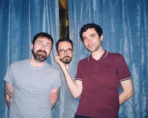 Trois hommes posent devant un rideau bleu. L'un d'eux, au centre, est un groupe musique espagnol année 2000.