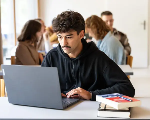 Ein junger Mann mit Schnurrbart konzentriert sich auf seinen Laptop, während er ein evangelisches Fernstudium Theologie absolviert.