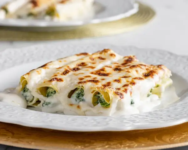 Cannelloni ricotta e spinaci fumanti, ricoperti da besciamella e formaggio fuso, serviti su un piatto bianco.