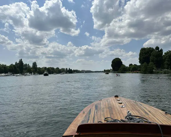 Blick vom Bug eines Holzbootes auf einen Fluss mit vielen Booten und grünen Ufern. Ein schöner Tag, fast wie ein Heidelberg See.