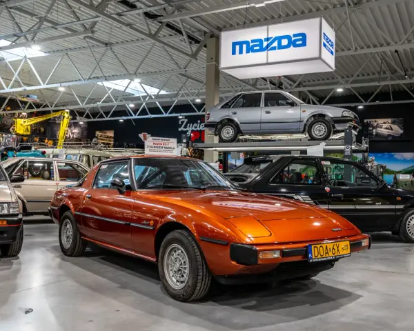 Pomarańczowa Mazda RX-7 błyszczy w muzeum samochodów, obok innych klasyków marki.