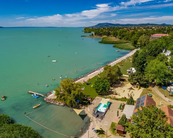 Słoneczny dzień na kempingu Balaton. Ludzie kąpią się w jeziorze i basenie, bawią na placu zabaw, a kampery stoją wśród drzew.
