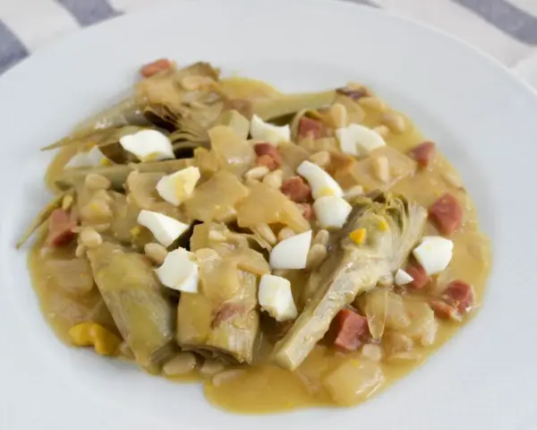 Plato de alcachofas con jamón, huevo duro y piñones, servido en una cazuela.