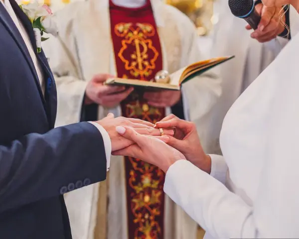 Ceremonia ślubna w kościele. Młoda para wymienia obrączki. W tle ksiądz z księgą. Warto wiedzieć, ile kosztuje ślub kościelny.