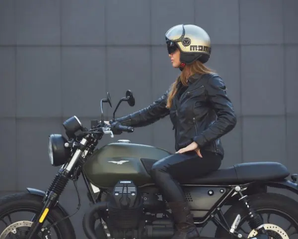 Donna in giacca di pelle e casco dorato, seduta su una moto verde. Non è chiaro dove si buttano i caschi da moto, ma questo è un ottimo esempio di come indossarli.