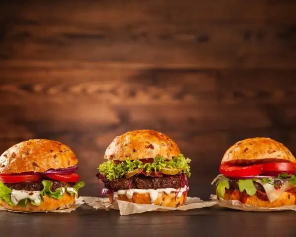 Trzy apetyczne burgery z różnymi dodatkami na drewnianym tle.