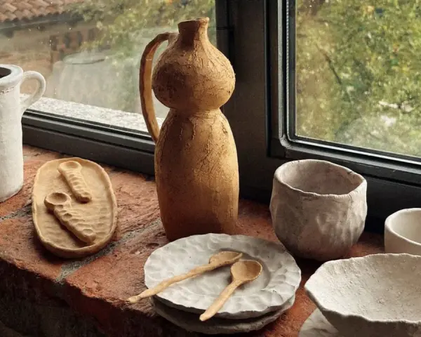 Ceramica fatta in casa: vasi, ciotole e cucchiaini grezzi, posati su un davanzale di mattoni, con vista su un giardino verde.