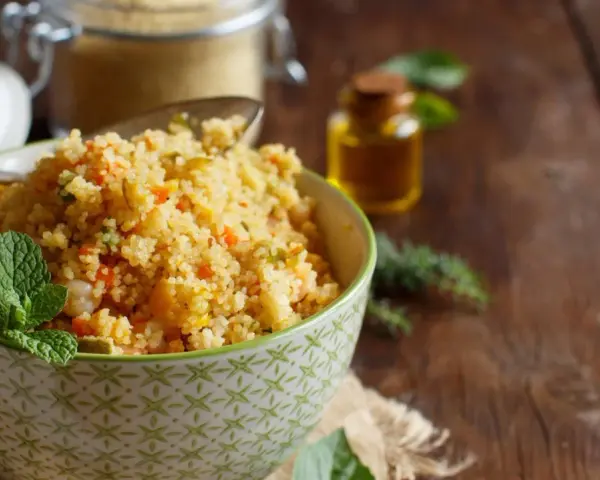 Schüssel mit buntem Couscous, garniert mit Minze. Daneben ein Glas Couscous und eine kleine Flasche Öl. Ein köstlicher Couscous-Baby-Genuss!
