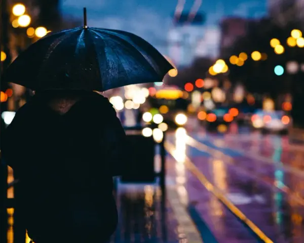 Person sous un parapluie noir, attendant sur un trottoir mouillé. Les lumières de la ville se reflètent sur la chaussée, créant de belles photos de pluie.