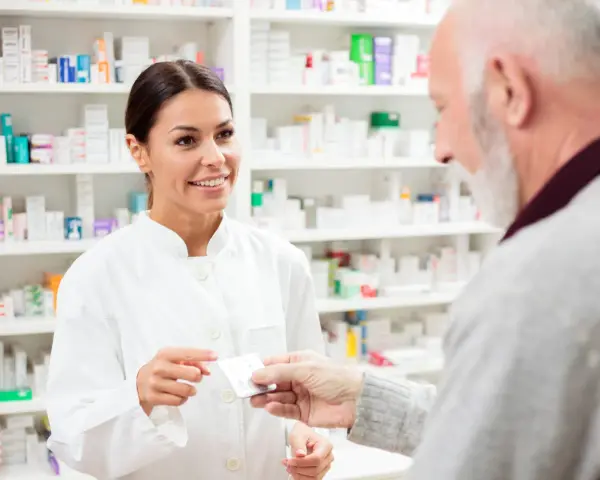 Uśmiechnięta farmaceutka podaje starszemu panu leki. W tle apteczne półki pełne leków, w tym poltixa.