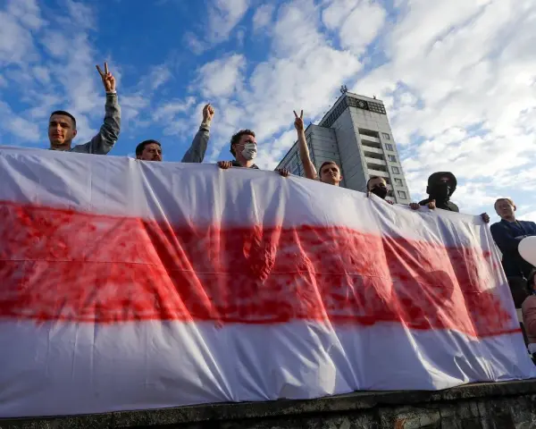 Ludzie trzymają biało-czerwoną flagę, symbol wolności Białorusi. Widać budynek w tle.