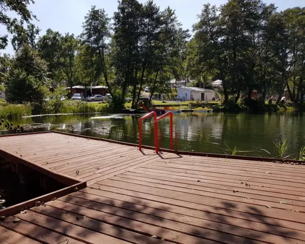 Drewniany pomost z czerwonymi poręczami nad jeziorem. W tle drzewa, budynki i samochody. Idealne miejsce na łagów camping.