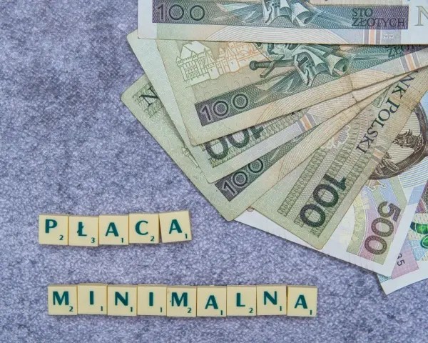 Płaca minimalna w Polsce – polskie banknoty o nominałach 100 i 500 zł ułożone na szarym tle, obok kafelki z literami tworzące napis "PŁACA MINIMALNA".