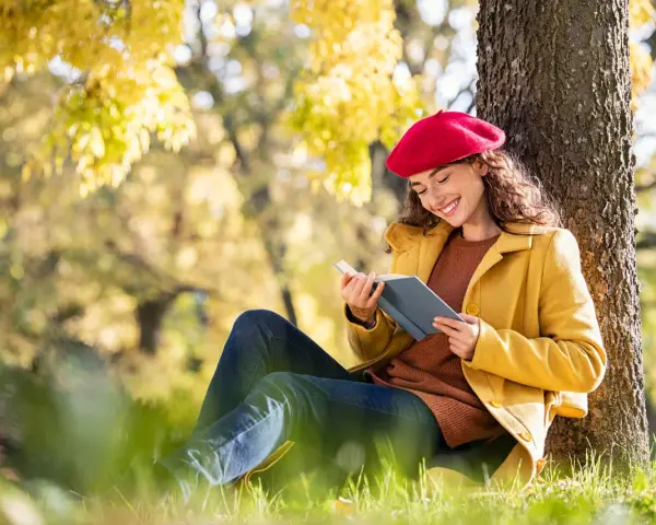 Mujer con boina roja lee un libro bajo un árbol, inspirada por los temas de cuentos del otoño.