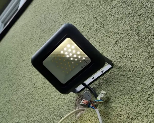 Czarna lampa led żarzy się po włączeniu, oświetlając zieloną, chropowatą ścianę. Widać przewody i złączki.