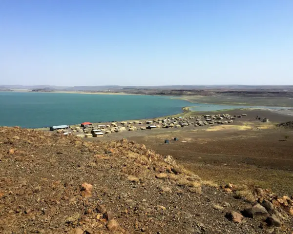 Widok na osadę nad turkusowym jeziorem Turkana. W oddali widać górzysty krajobraz.
