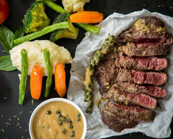 Succulenta costata di manzo affettata, servita con purè, verdure miste e salsa al pepe verde.
